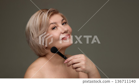 Mature woman posing with makeup brush near chin. Mature woman posing with makeup brush near chin. 136711140
