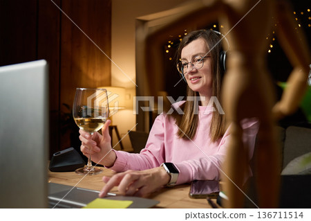 Woman wearing headphones uses laptop at desk while holding wine glass in home interior Woman wearing headphones uses laptop at desk while holding wine glass in home interior 136711514