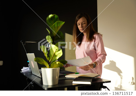 Woman holding paper documents while standing at desk with laptop Woman holding paper documents while standing at desk with laptop 136711517