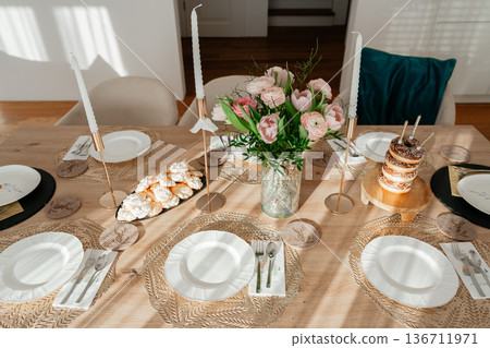 Beautifully arranged dining table with plates, candles and fresh flowers. Warm sunlight creates inviting atmosphere perfect for restaurant, hospitality and event advertising 136711971