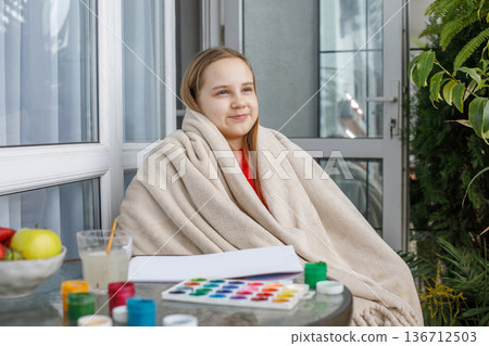 Cheerful child in a warm fleece blanket relaxing on the balcony on cool spring day. 136712503
