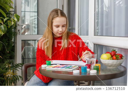 Cheerful young girl in red warm sweater drawing in a sketchbook while sitting on the balcony Cheerful young girl in red warm sweater drawing in a sketchbook while sitting on the balcony 136712510