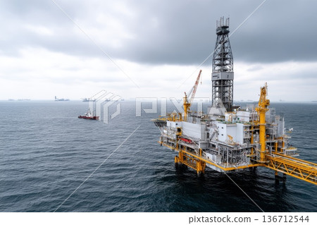 Offshore construction platform for oil and gas production in the ocean, with a support vessel visible and a cloudy sky creating a dramatic backdrop Offshore construction platform for oil and gas production in the ocean, with a support vessel visible and a cloudy sky creating a dramatic backdrop 136712544