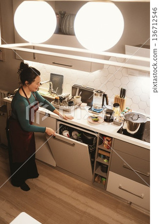 Woman in a kitchen loading a dishwasher with a smile, highlighting a clean and organized cooking space. Light from overhead enhances the workspace, creating a cozy atmosphere. Woman in a kitchen loading a dishwasher with a smile, highlighting a clean and organized cooking space. Light from overhead enhances the workspace, creating a cozy atmosphere. 136712546