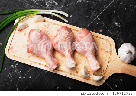 Top view of three raw chicken drumsticks on a wooden cutting board with garlic cloves, green onions, and pink salt, set against a dark marble background. High quality photo Top view of three raw chicken drumsticks on a wooden cutting board with garlic cloves, green onions, and pink salt, set against a dark marble background. High quality photo 136712755