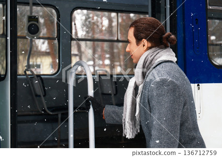 Smiling man in a grey coat boarding a tram during a winter snowfall. He holds the handrail while entering public transport in cold weather. High quality photo Smiling man in a grey coat boarding a tram during a winter snowfall. He holds the handrail while entering public transport in cold weather. High quality photo 136712759