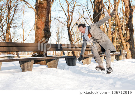 Man slipping and falling on snow near a bench in a winter park. Young guy losing balance on slippery ice during a sunny cold day. Winter accident and safety concept. High quality photo 136712919