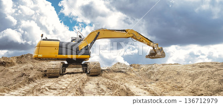 An excavator operates on a construction site, moving dirt with its bucket while clouds fill the sky above. The equipment is in action during the day 136712979