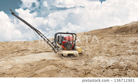 A vibratory plate compactor works on a sandy area at a construction site. Preparing the foundation for construction A vibratory plate compactor works on a sandy area at a construction site. Preparing the foundation for construction 136712981