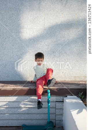Young Child Sitting Thoughtfully On A Scooter Bench 136713424
