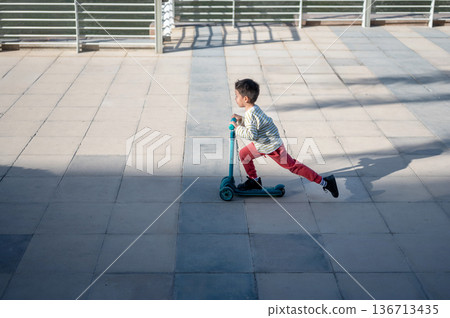 Child Enthusiastically Riding A Scooter By The Waterside Path Child Enthusiastically Riding A Scooter By The Waterside Path 136713435
