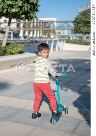 Young Boy Riding A Scooter In Playful Outdoor Setting With Joy. 136713437