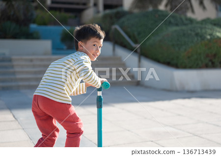 Young Boy Riding A Scooter In Playful Outdoor Setting With Joy. 136713439