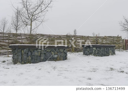Snow-covered benches in a nature park. Benches coated in snow with rustic wooden fencing in the background. Concept of winter and winter nature. Snow-covered benches in a nature park. Benches coated in snow with rustic wooden fencing in the background. Concept of winter and winter nature. 136713499