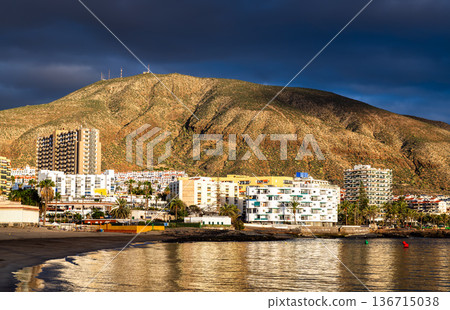 View of Playa de Los Cristianos beach and Mount Guaza in Tenerife, Canary Islands, Spain. Features the sandy beach, calm ocean water, and resort buildings illuminated at sunset. View of Playa de Los Cristianos beach and Mount Guaza in Tenerife, Canary Islands, Spain. Features the sandy beach, calm ocean water, and resort buildings illuminated at sunset. 136715038