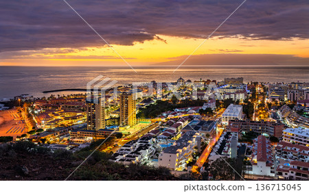 The resort town of Los Cristianos illuminated at sunset in Tenerife, Canary Islands, Spain. The view from Mount Chayofita volcano overlooks the Atlantic Ocean with La Gomera island on the horizon 136715045