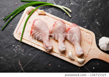 Top view of three raw chicken drumsticks on a wooden cutting board with green onions, garlic cloves, and pink salt on a dark marble background. High quality photo 136715325