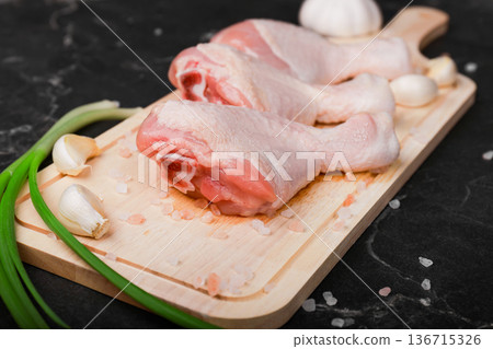 Raw chicken drumsticks on a wooden board with garlic, green onions, and pink salt on a dark marble background prepared for cooking. High quality photo 136715326