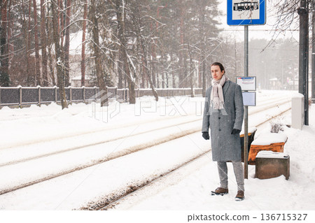 Man in a grey coat waits for a tram at a snowy stop on a cold winter day. He stands by the tracks looking for transport, potentially late for work during a snowstorm. High quality photo 136715327