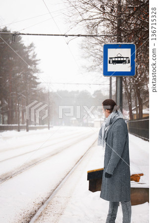 Man in a grey coat stands at a snowy tram stop, looking down the empty tracks. He waits impatiently for public transport on a cold winter day, fearing he is late. High quality photo 136715328