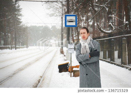 Cold man in a grey coat standing at a snowy tram stop with crossed arms. He looks impatient and frozen while waiting for public transport during winter weather. High quality photo Cold man in a grey coat standing at a snowy tram stop with crossed arms. He looks impatient and frozen while waiting for public transport during winter weather. High quality photo 136715329