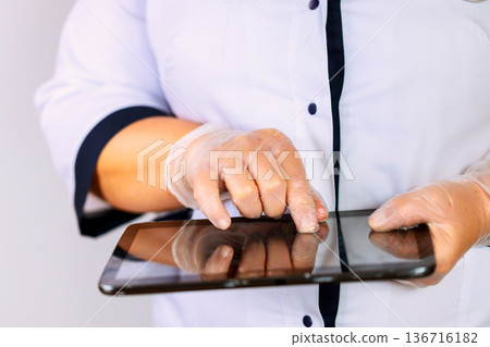 Nurse wearing gloves is using tablet with one hand while other hand touches screen in medical office 136716182