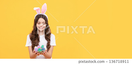 smiling child in bunny ears hold eggs on yellow background. Easter child horizontal poster. Web banner header of bunny kid, copy space. 136718911