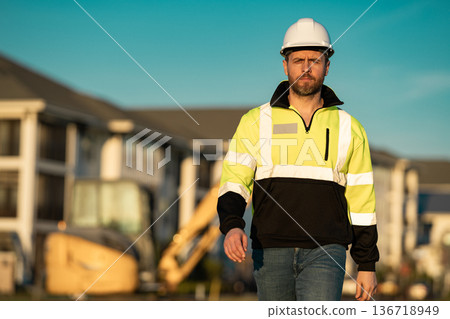 Caucasian man, construction worker in helmet at construction site. Industry engineer worker in hardhat near bulldozer or digger tractor. Concept of construction industry. Construction site manager. 136718949