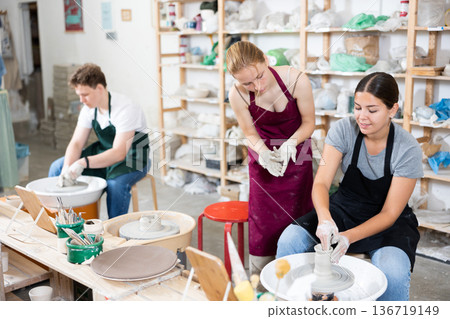 Female potter teaches young apprentices how to make pottery on potter wheel in workshop Female potter teaches young apprentices how to make pottery on potter wheel in workshop 136719149