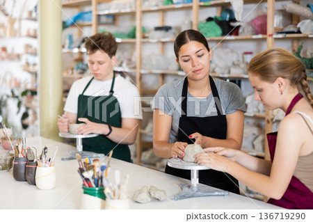 Young woman teacher helps teenagers boy and girl to sculpt ceramic product Young woman teacher helps teenagers boy and girl to sculpt ceramic product 136719290