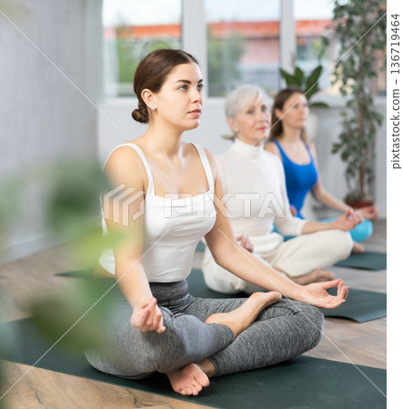 Calm women of different ages meditating in Lotus Pose or Padmasana in beginning yoga training together in cozy gym studio Calm women of different ages meditating in Lotus Pose or Padmasana in beginning yoga training together in cozy gym studio 136719464