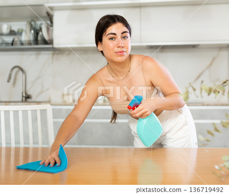 Positive young woman cleaning the surface of kitchen table Positive young woman cleaning the surface of kitchen table 136719492