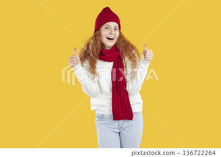 Happy friendly girl in red hat, scarf showing thumbs up looking at camera on yellow background. 136722264