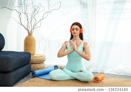 Woman sits on a mat with headphones, practicing yoga focus Woman sits on a mat with headphones, practicing yoga focus 136724180