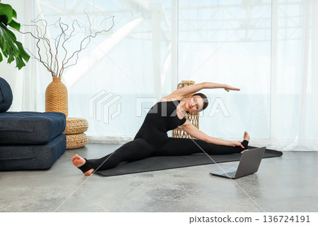 A woman performs yoga stretches at home with a laptop A woman performs yoga stretches at home with a laptop 136724191