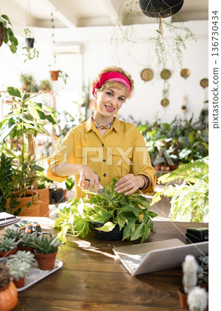 A smiling woman prunes a houseplant in a bright, airy greenhouse, surrounded by lush greenery A smiling woman prunes a houseplant in a bright, airy greenhouse, surrounded by lush greenery 136730434