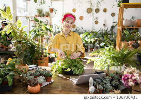 A woman smiles while trimming a houseplant in a bright, plant-filled greenhouse setting A woman smiles while trimming a houseplant in a bright, plant-filled greenhouse setting 136730435