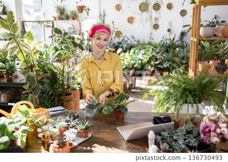 A woman in a yellow shirt smiles while tending to plants in a bright, inviting shop 136730493