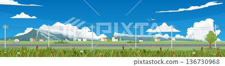 Landscape of an empty asphalt road. Cutting through wide fields lined with power poles. Small village sits on hill with meadows and flowers in foreground. Mountains in the background under a blue sky. 136730968