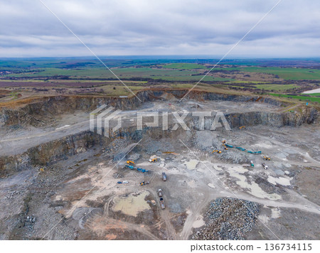 Large excavation site showing heavy machinery working on a quarry surrounded by green fields and cloudy skies in the background Large excavation site showing heavy machinery working on a quarry surrounded by green fields and cloudy skies in the background 136734115