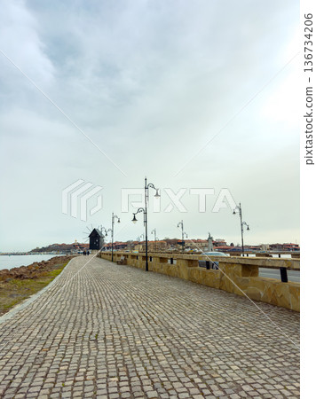 Visitors walk along a cobblestone path beside the water at a harbor while old buildings and a windmill are visible in the distance during a cloudy day Visitors walk along a cobblestone path beside the water at a harbor while old buildings and a windmill are visible in the distance during a cloudy day 136734206