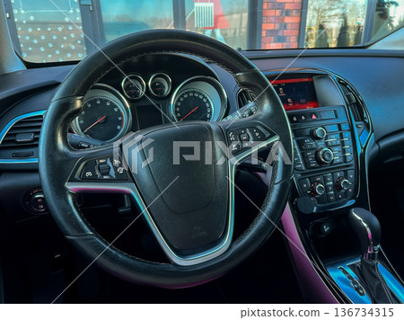 Interior view of a car dashboard showing steering wheel and controls in a vehicle parked near a building during daylight Interior view of a car dashboard showing steering wheel and controls in a vehicle parked near a building during daylight 136734315