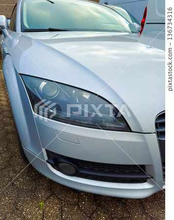 Silver car parked in a lot with close-up view of the front, showing details of the headlights and grill in a typical urban setting during daylight Silver car parked in a lot with close-up view of the front, showing details of the headlights and grill in a typical urban setting during daylight 136734316