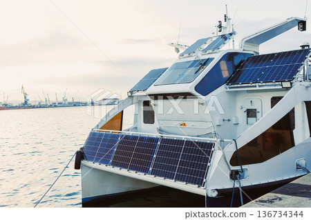 Solar powered boat docked at the harbor during early morning with city skyline in the background and calm water reflecting the light Solar powered boat docked at the harbor during early morning with city skyline in the background and calm water reflecting the light 136734344