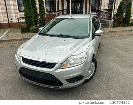 Silver car parked on a street near a building with green trees in the background on a cloudy day Silver car parked on a street near a building with green trees in the background on a cloudy day 136734352