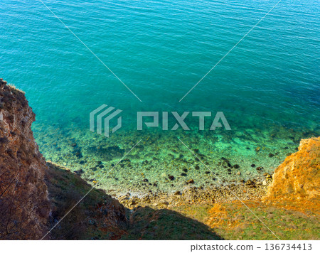 Water and rocks meet at a clear shoreline with green and blue colors under bright sunlight during the day Water and rocks meet at a clear shoreline with green and blue colors under bright sunlight during the day 136734413