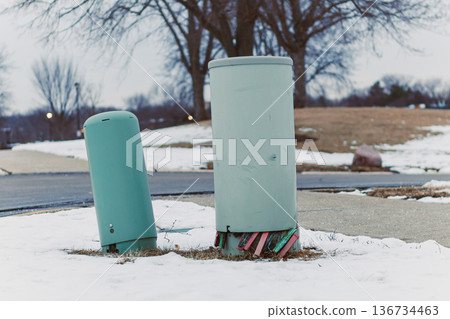 Two green utility poles stand on the edge of a snow-covered area near a sidewalk in a suburban park during winter 136734463