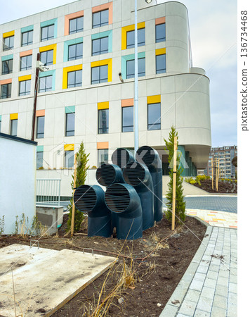 Multiple ventilation pipes stand near a modern building with colorful panels in a city area on a cloudy day Multiple ventilation pipes stand near a modern building with colorful panels in a city area on a cloudy day 136734468