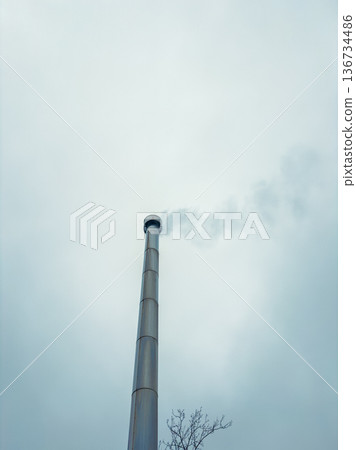 Smoke rises from a tall industrial chimney under a cloudy sky in an urban area during the afternoon hours in late autumn Smoke rises from a tall industrial chimney under a cloudy sky in an urban area during the afternoon hours in late autumn 136734486