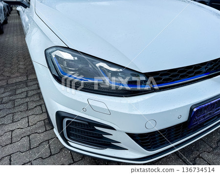 Close view of a white car parked in a lot with wet pavement on a cloudy day showcasing modern design features and lighting Close view of a white car parked in a lot with wet pavement on a cloudy day showcasing modern design features and lighting 136734514